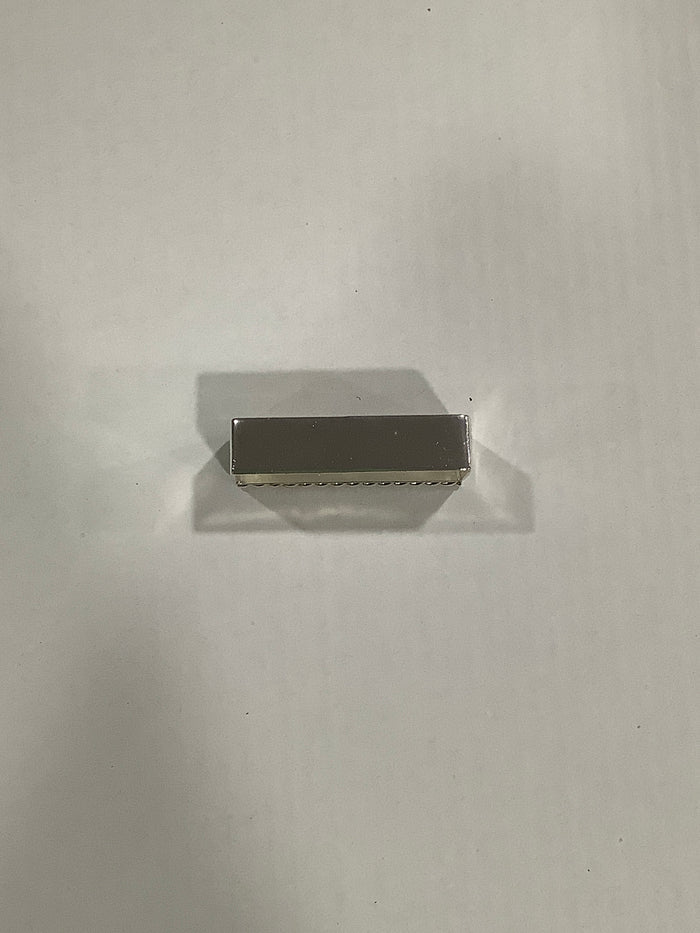 A small rectangular metallic object with a brushed silver finish, positioned horizontally on a plain light gray surface with a soft shadow.