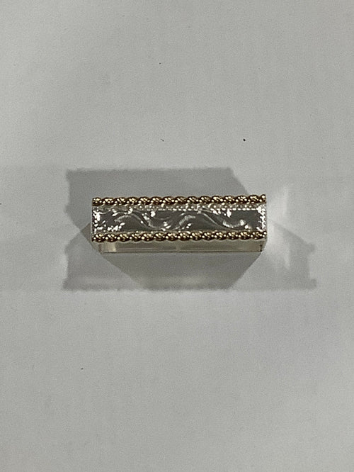A small rectangular silver-toned metal charm with an engraved floral pattern and a textured gold-toned beaded border, shown on a plain light gray background.