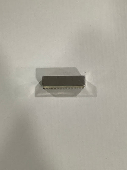 A small rectangular metallic object with a brushed silver finish, positioned horizontally on a plain light gray surface with a soft shadow.